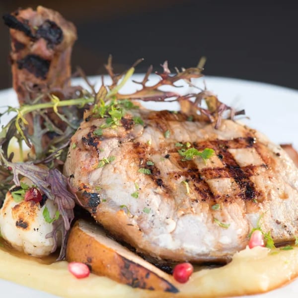 Grilled pork chop with mashed potatoes, roasted pears, and greens on a white plate.