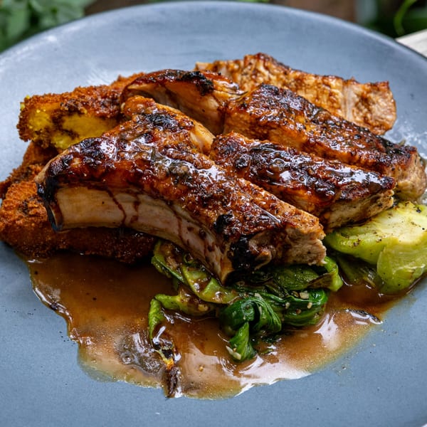 Glazed ribs with crispy fried plantains and green vegetables in a rich sauce on a blue plate.