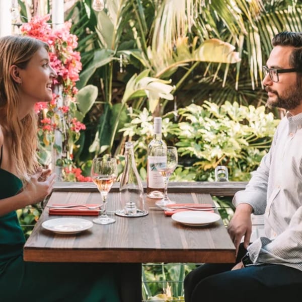 A couple enjoying a meal and drinks on an outdoor patio surrounded by lush greenery and pink flowers.