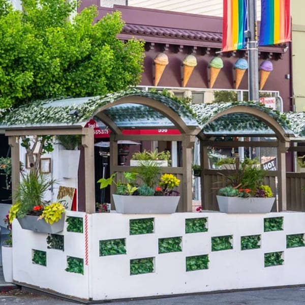 Outdoor dining patio with planters, green walls, and a rainbow flag in the background.